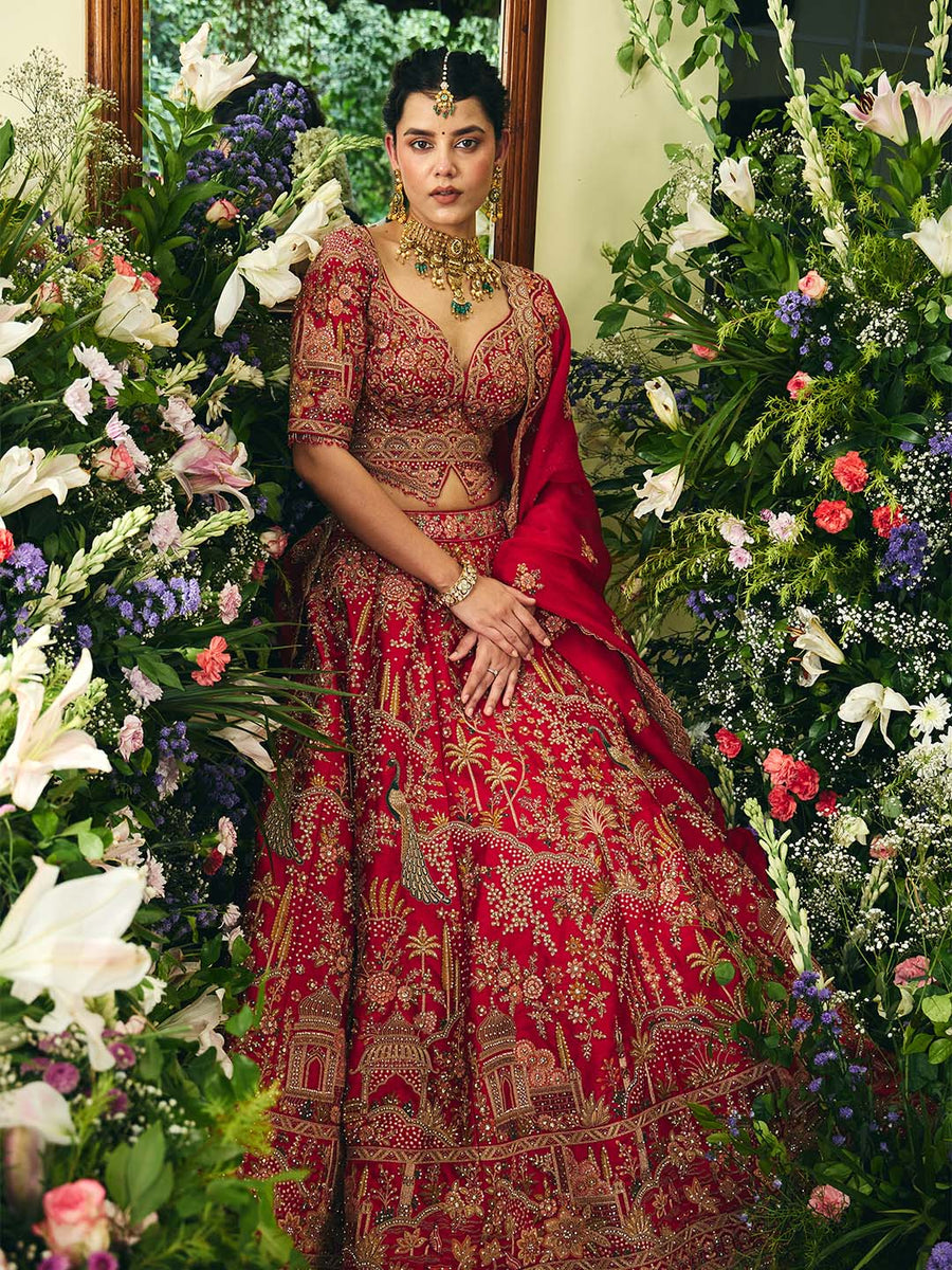 Woman dressed in a red embroidered lehenga set posing with matching dupatta and detailed traditional embroidery in a floral setting