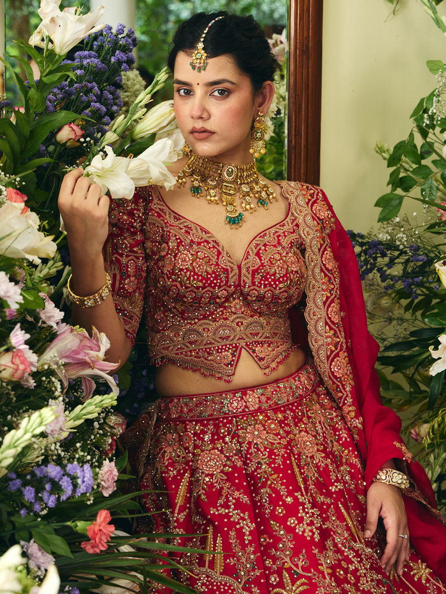 Close-up of a woman in red bridal lehenga showcasing detailed zari, zardozi, and bead embroidery holding white flowers among greenery
