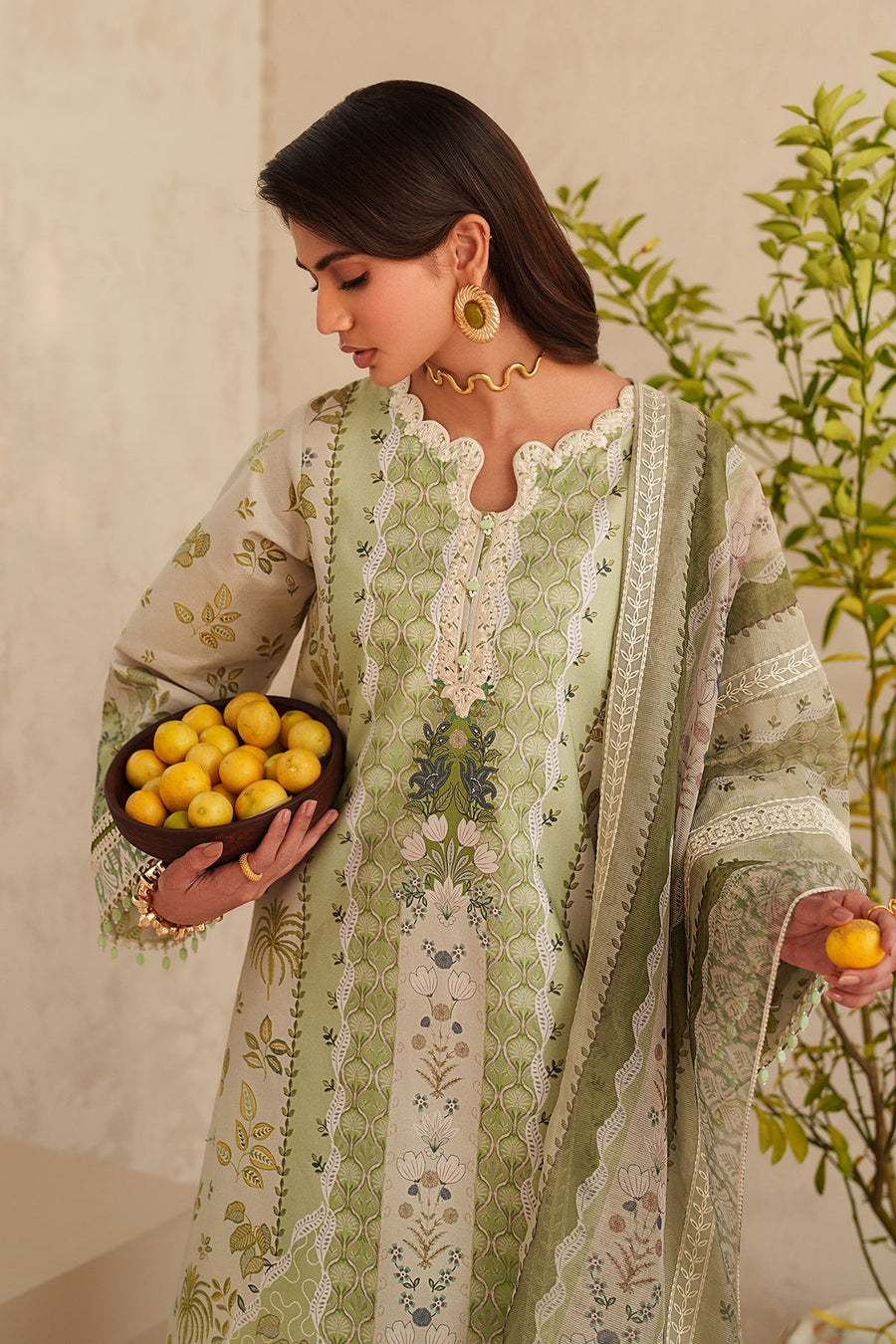 Woman standing with elegant embroidered lawn dupatta draped over shoulders, holding lemons in both hands, decorative setting
