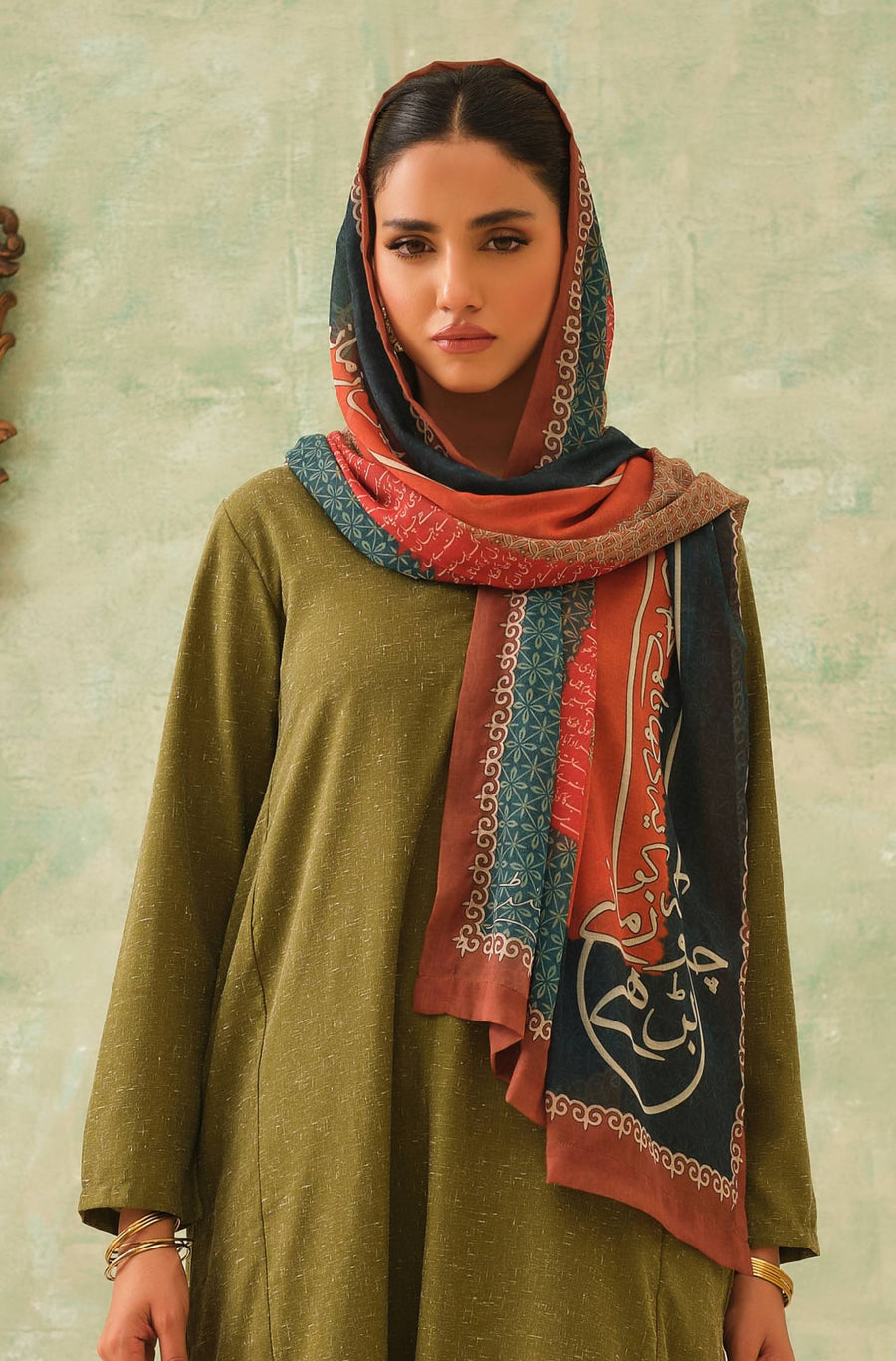 Woman wearing an embroidered scarf with vibrant teal, burnt orange, and beige Urdu calligraphy print paired with olive green traditional wear.