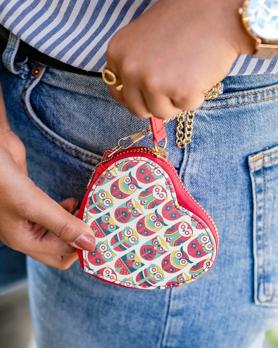 Close-up view of a woman’s hand holding a heart-shaped bag charm with colorful owl prints and gold chain, attached to a belt loop on denim jeans