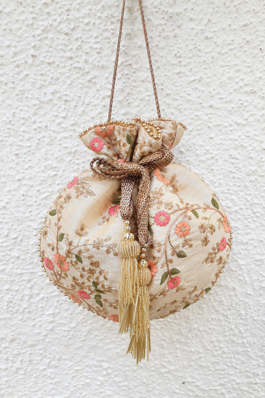 Cream round potli bag with floral embroidery and gold beaded tassels hanging against textured wall