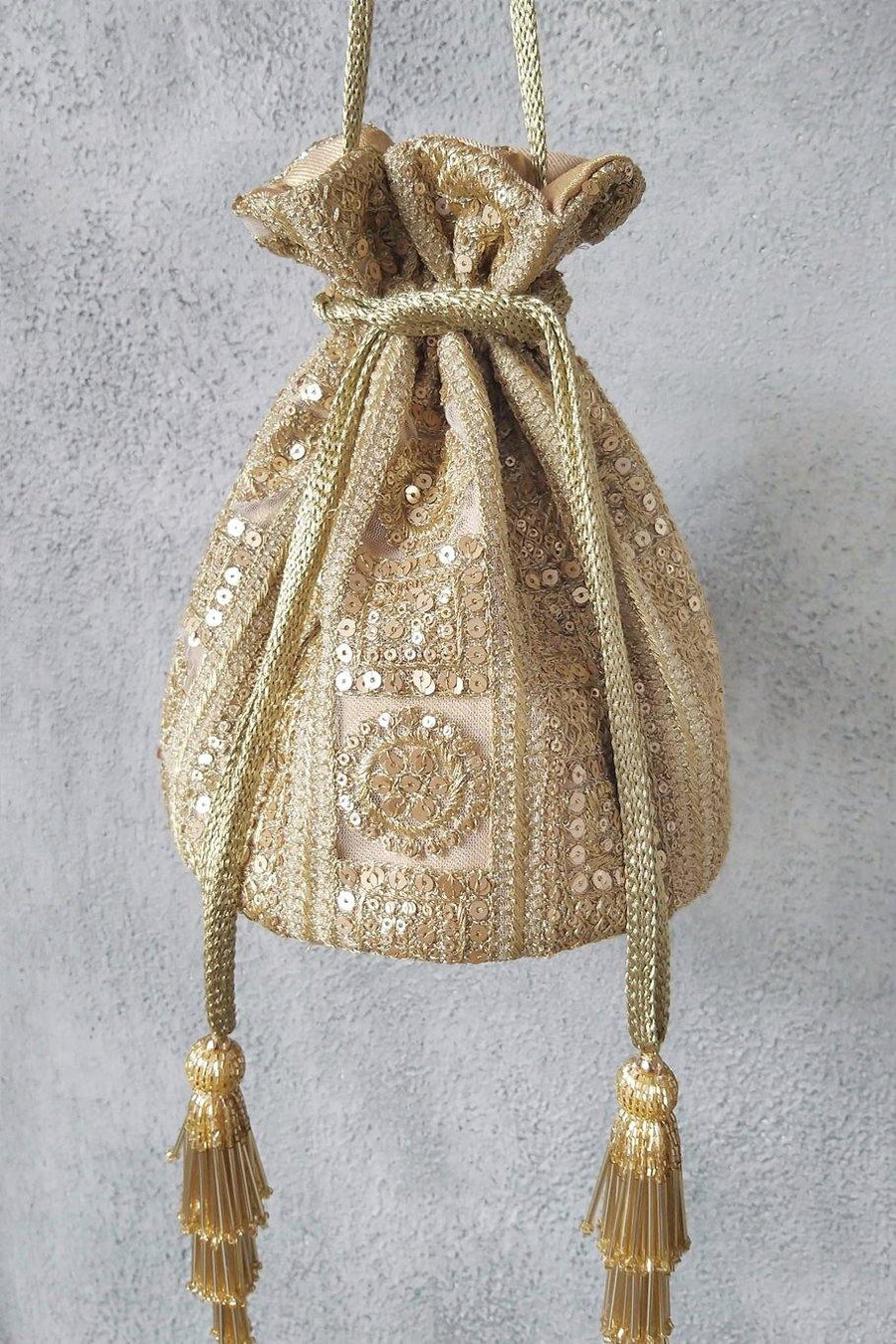 Gold potli bag with intricate sequin and zari embroidery on net fabric, featuring gold braided drawstrings and tassels, hanging against a textured background