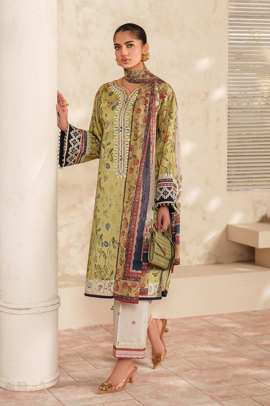 Woman posing near a pillar wearing floral embroidered lawn shirt, white pants, and a matching floral dupatta draped over her shoulder