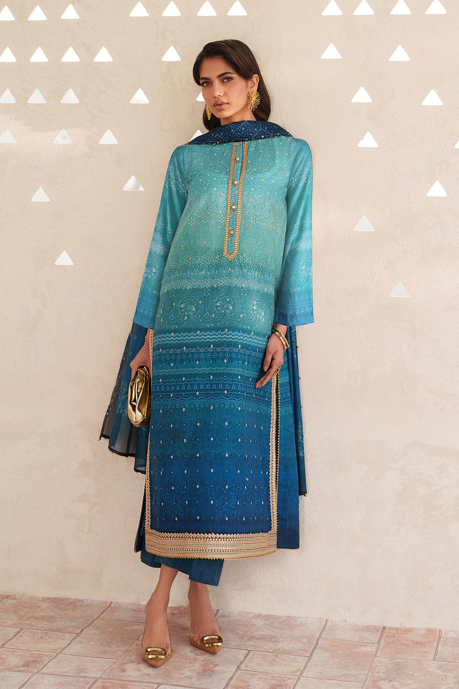 Woman wearing a teal to navy blue ombre embroidered lawn suit with gold detailing, holding a gold clutch and wearing gold earrings and heels