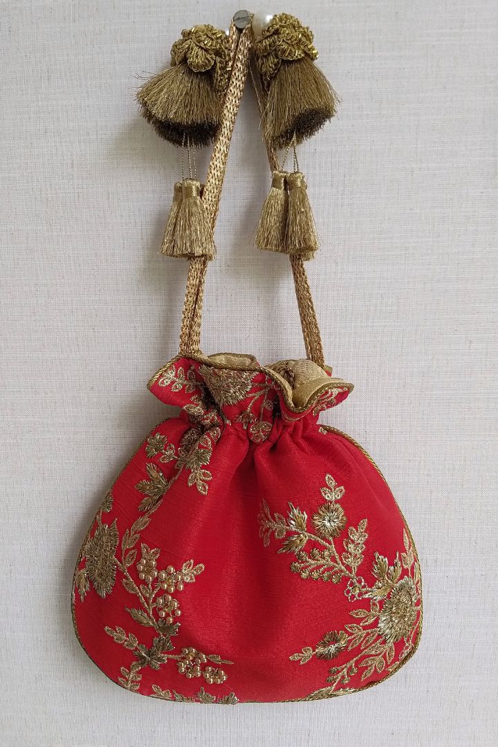 Red silk potli bag with gold floral embroidery and decorative tassels hanging against a neutral background