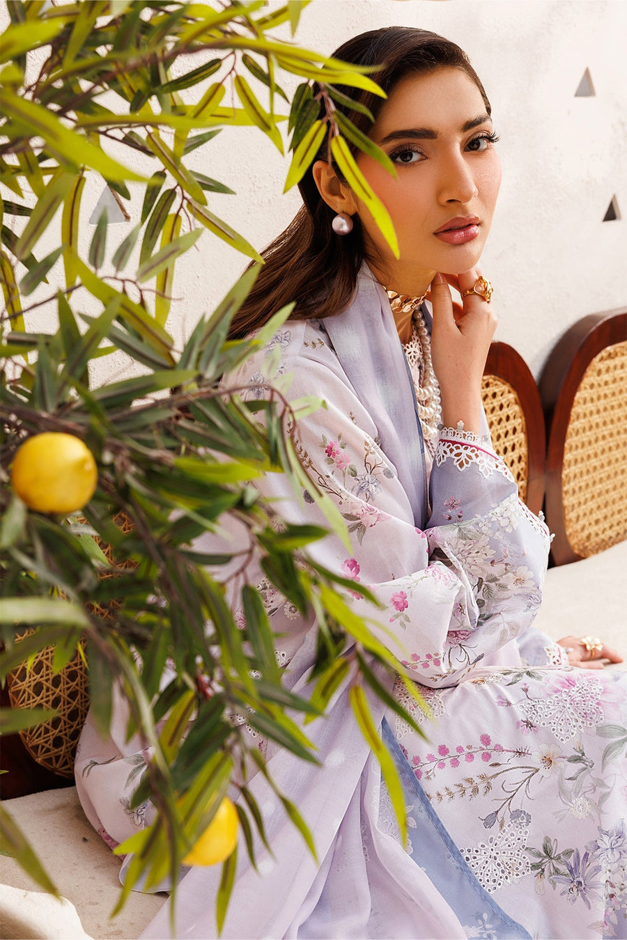 Full body shot of woman in lavender floral lawn suit posing seated on a wicker chair, offering a clear view of dress and accessories.