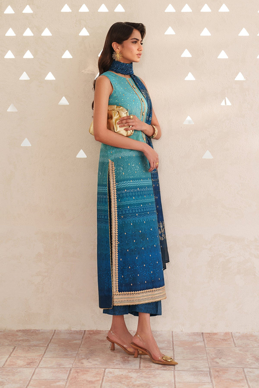 Woman sitting on a wooden bench wearing the blue embroidered lawn suit with detailed dupatta, styled with gold clutch and jewelry