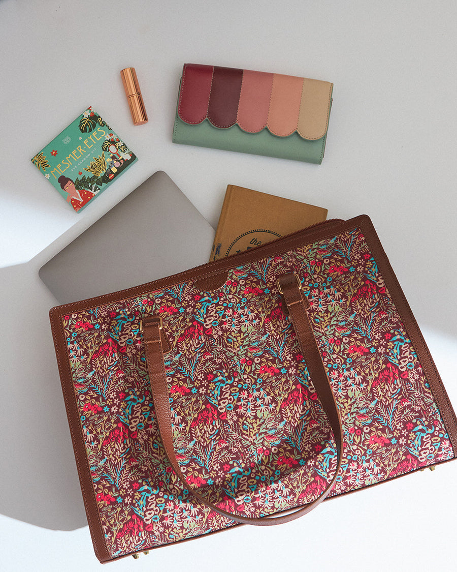 Overhead view of floral patterned tote bag with brown leather handles alongside a colorful wallet, makeup, and notebook