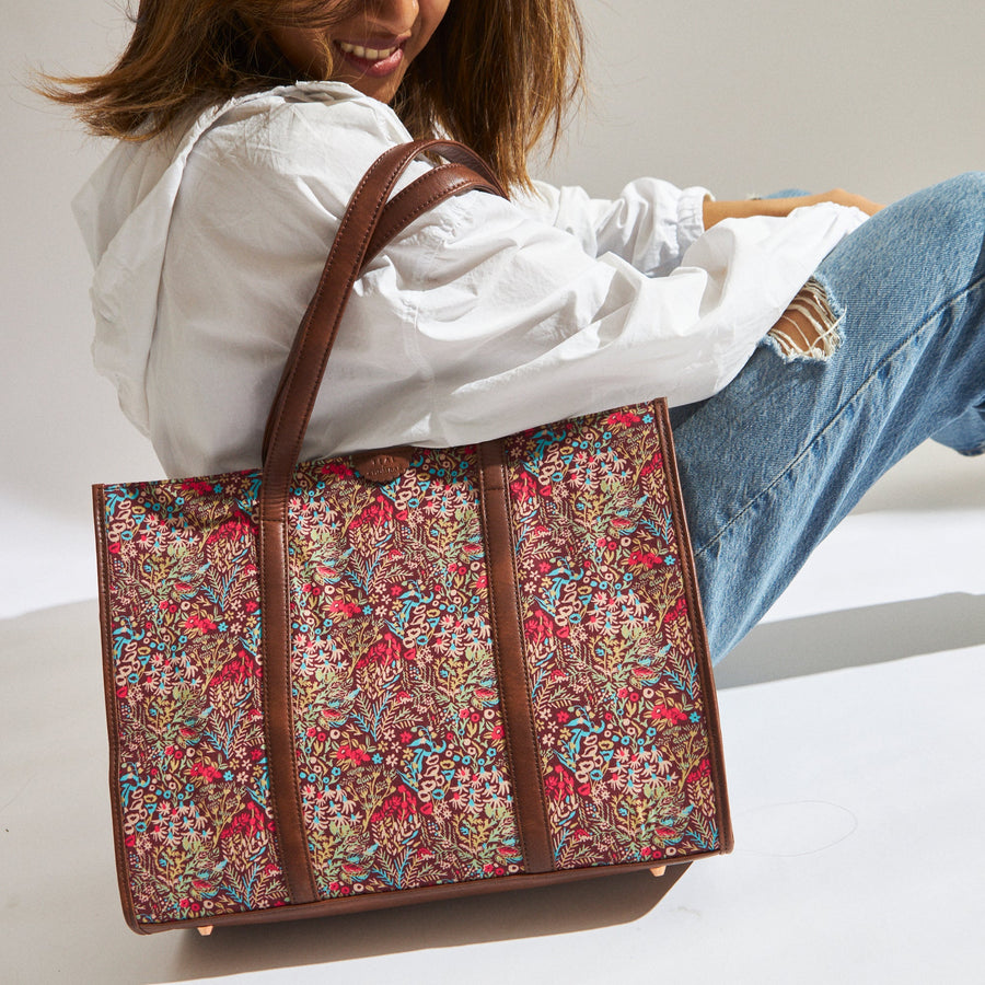 Woman holding vibrant floral canvas tote bag with brown vegan leather straps and accents, wearing ripped blue jeans and white jacket