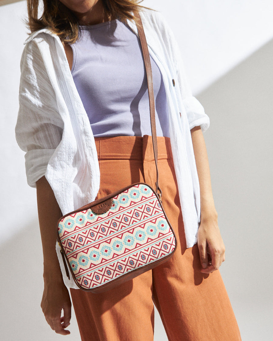 Woman wearing an Aztec patterned rectangular crossbody bag with leather strap, paired with a white shirt, lavender top, and rust orange pants.