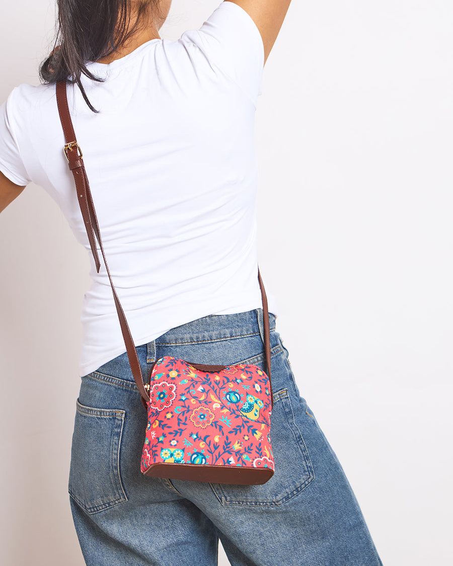 Woman wearing a floral patterned coral sling bag with an adjustable brown strap, paired with blue jeans and a white t-shirt, shown from the back