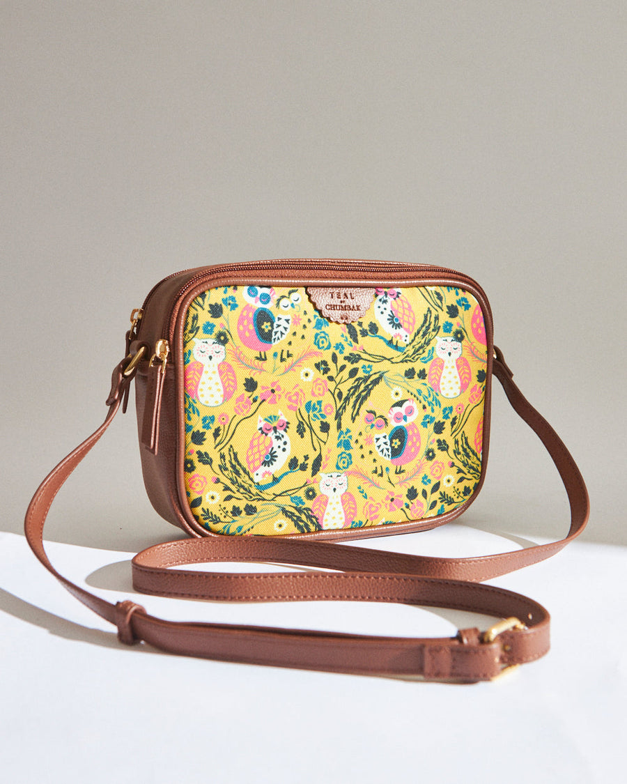 Brown leather box sling bag with vibrant yellow front panel featuring colorful owl and floral print, adjustable strap laid out on white surface.