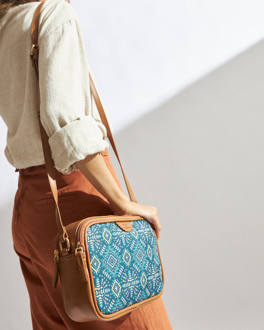 Woman wearing a teal Aztec print box sling bag with tan leather trim and adjustable strap paired with cream top and rust pants