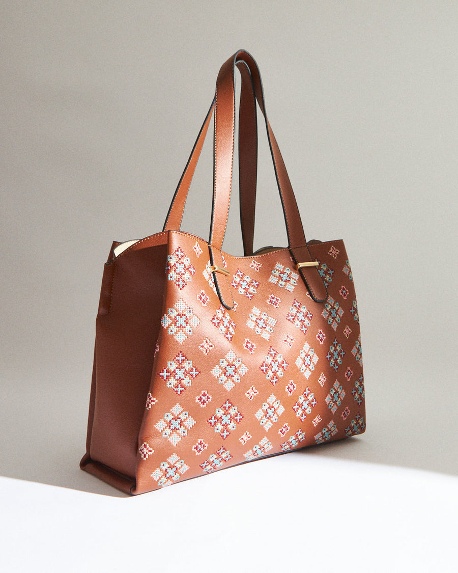 Side angled view of a spacious tan faux leather tote bag with cross stitch detail and structured shape
