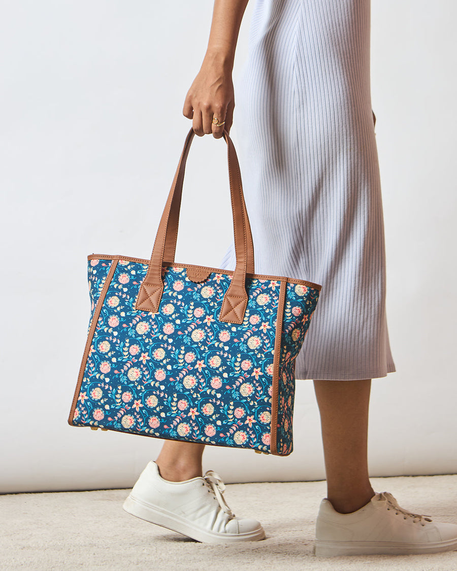 Woman holding a blue floral patterned tote bag with brown leather handles and trim, wearing a white ribbed skirt and white sneakers