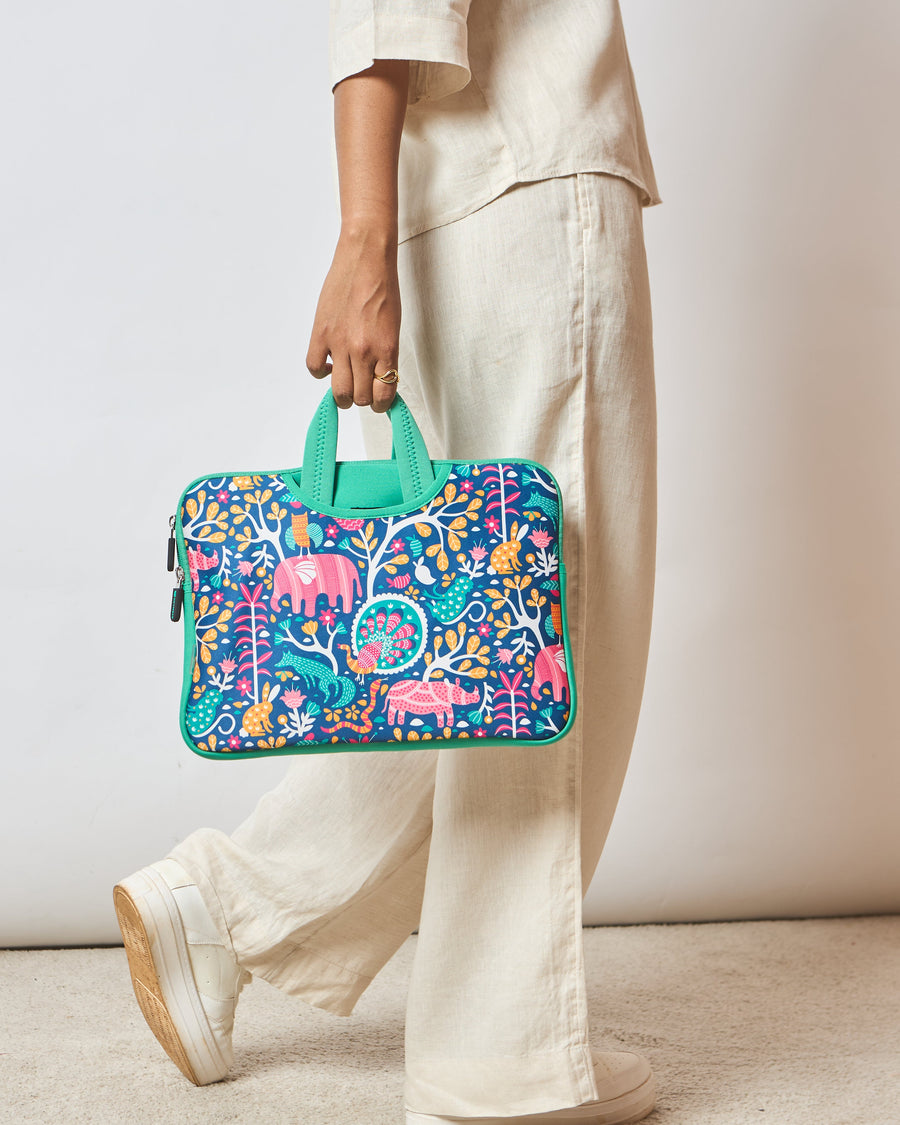 Woman holding a colorful Sundarbans laptop sleeve with vibrant Gondh art on a blue background, featuring pink, orange, and green jungle animals and plants, carrying it by mint green handles, dressed in beige clothing and white sneakers.