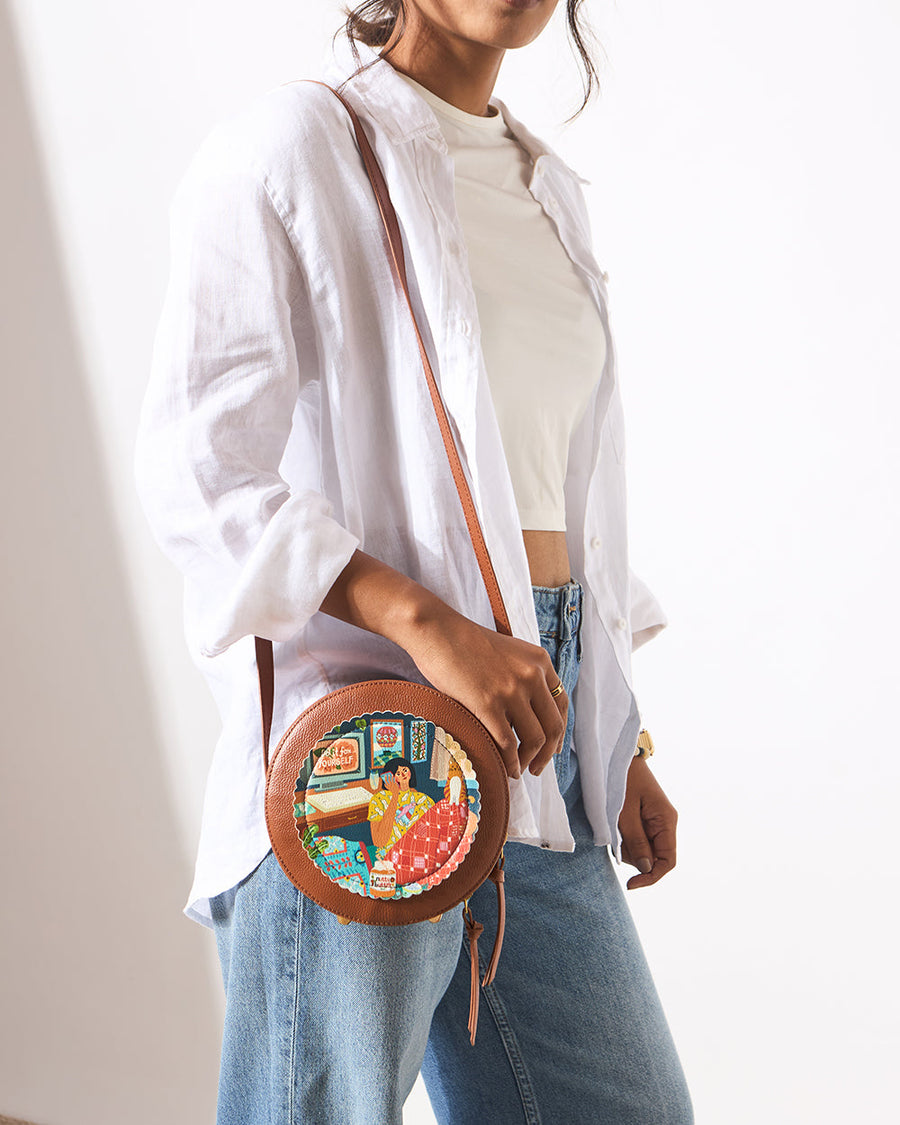 Woman wearing a white blouse and jeans carrying a brown round leather crossbody bag with a colorful artistic print and adjustable strap