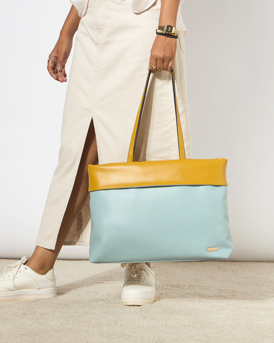 Woman holding a two-tone tote bag with mustard yellow top and light blue body, paired with white sneakers and cream skirt