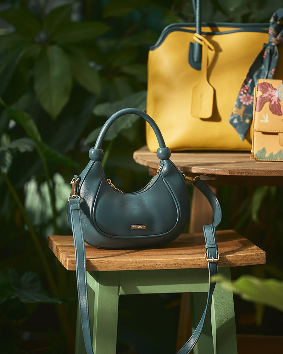 Deep teal vegan leather crescent-shaped handbag on green stool with yellow handbag background