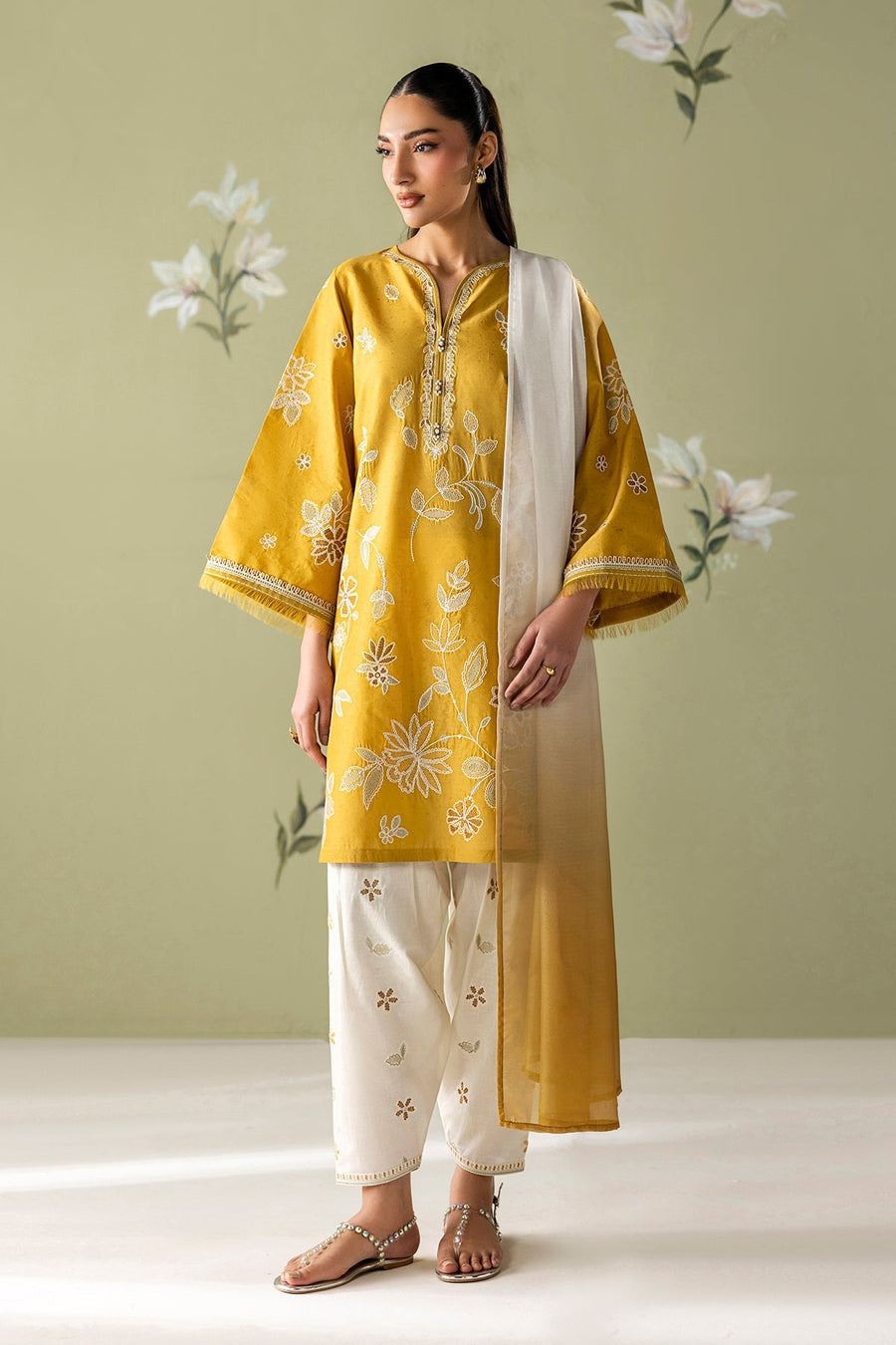 Model wearing embroidered mustard yellow lawn shirt with white floral patterns paired with white embroidered pants and white to mustard ombré dupatta against a floral green background