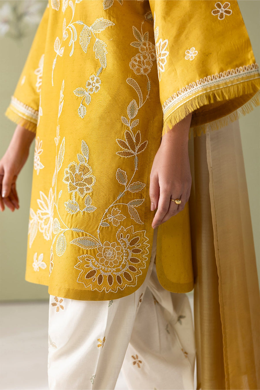 Detailed close-up of mustard yellow shirt's floral embroidery and fringe sleeve border with white floral embroidery on pants