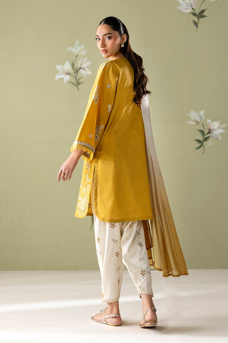 Back view of model in mustard yellow embroidered lawn shirt with white embroidery on sleeves and white embroidered pants, holding an ombré dupatta