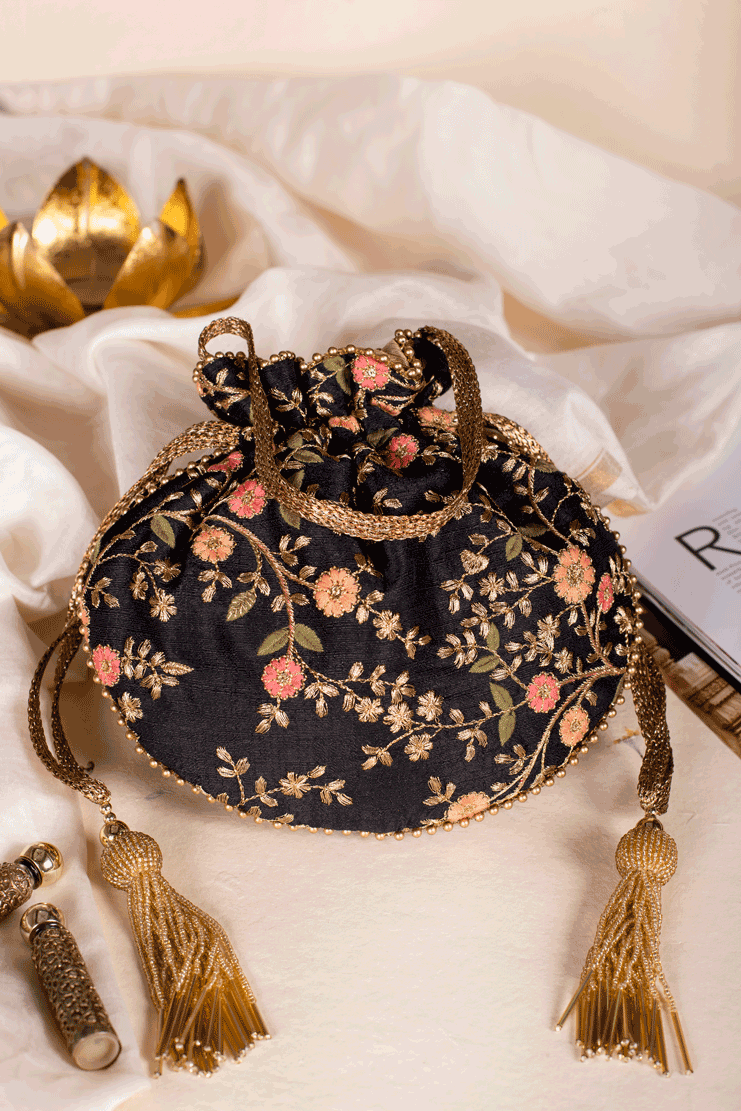 Black potli bag with intricate gold and blush floral embroidery, golden drawstring closure, and decorative tassels displayed on ivory fabric