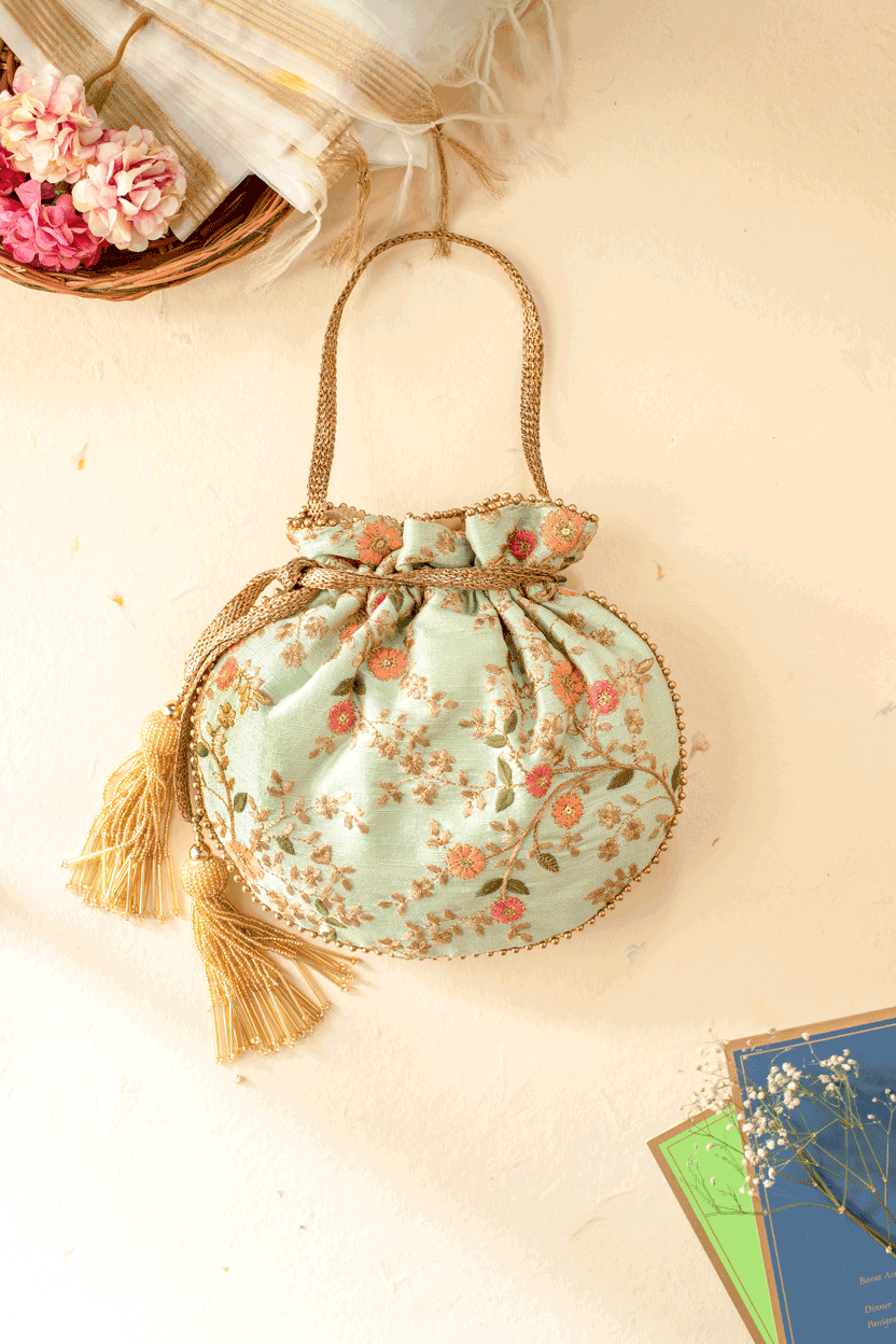 Mint-colored potli bag with intricate pastel floral embroidery, gold beaded piping, and tassel drawstrings hanging against a cream wall background