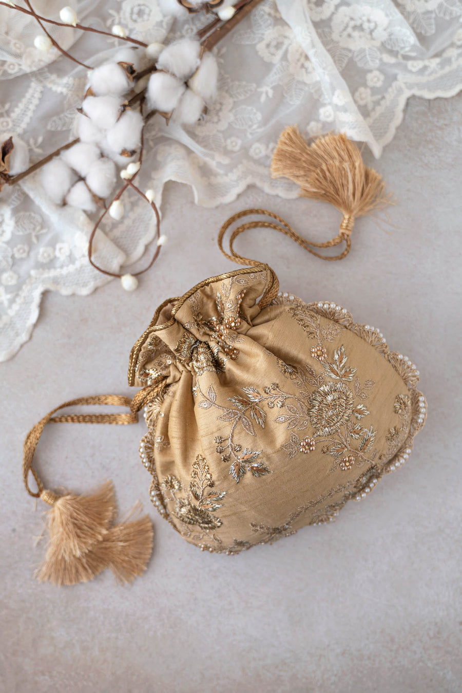 Gold potli bag with intricate floral embroidery and pearl accents, resting on lace fabric background with cotton branches