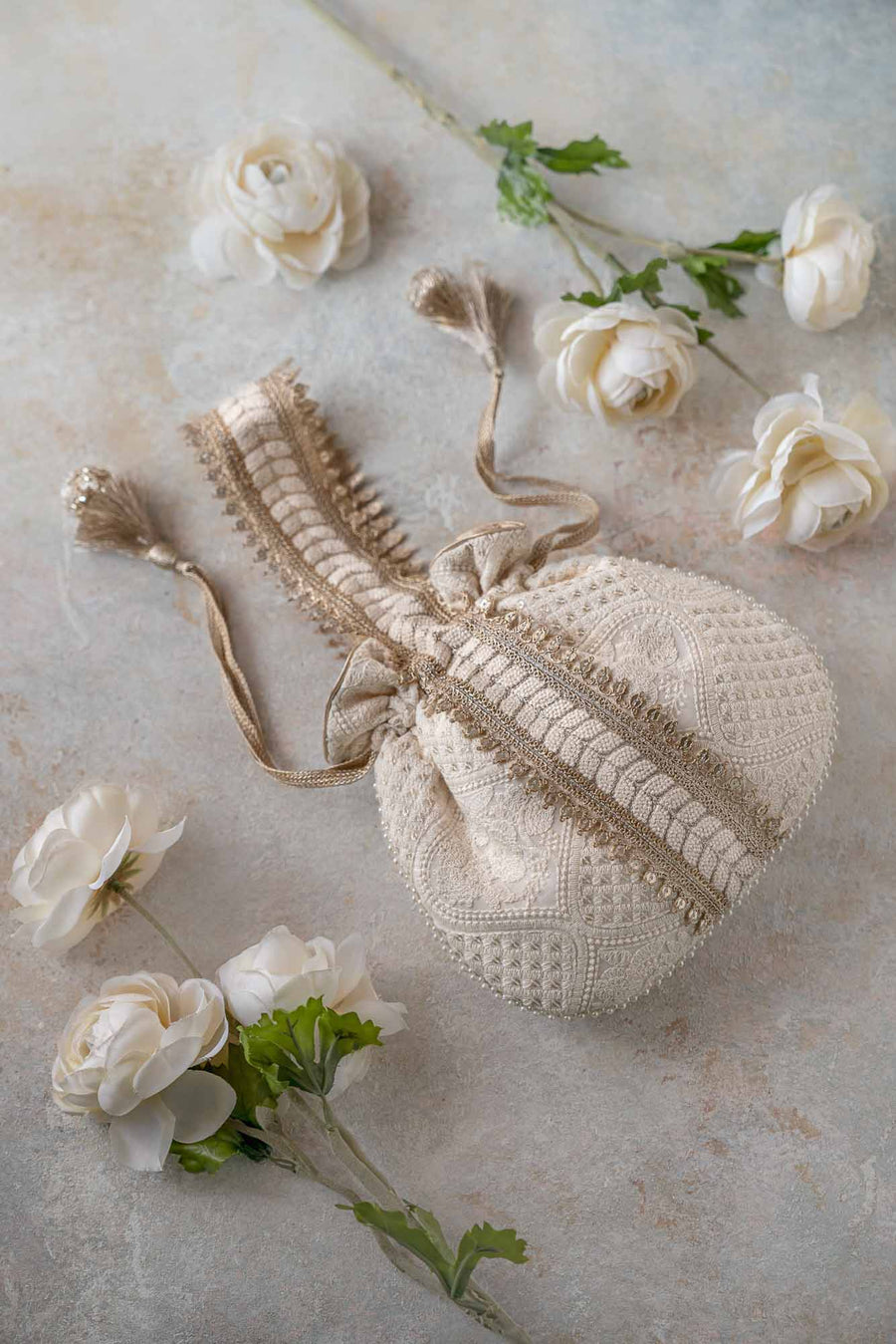 Off-white Mughal potli bag featuring intricate embroidery with gold zari detail, placed on a neutral stone surface with white floral accents.