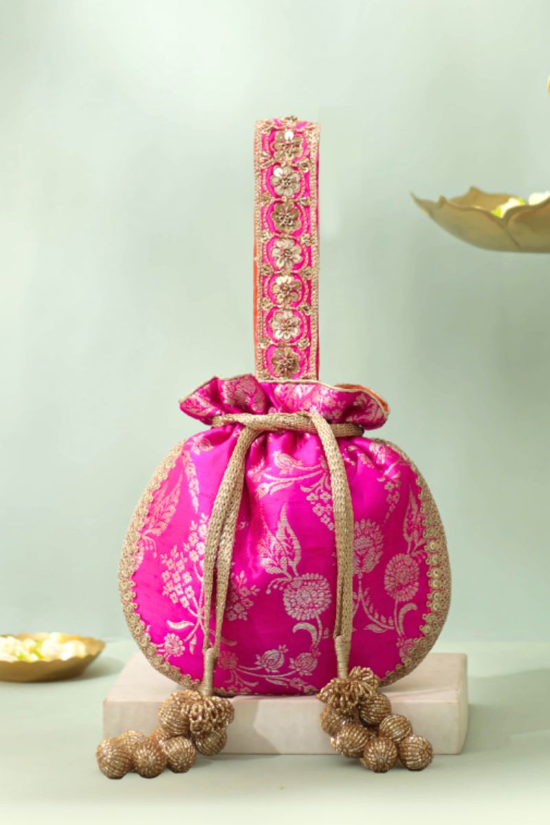 Pink silk potli bag with golden floral embroidery, decorated handle, and golden tassels on a neutral background