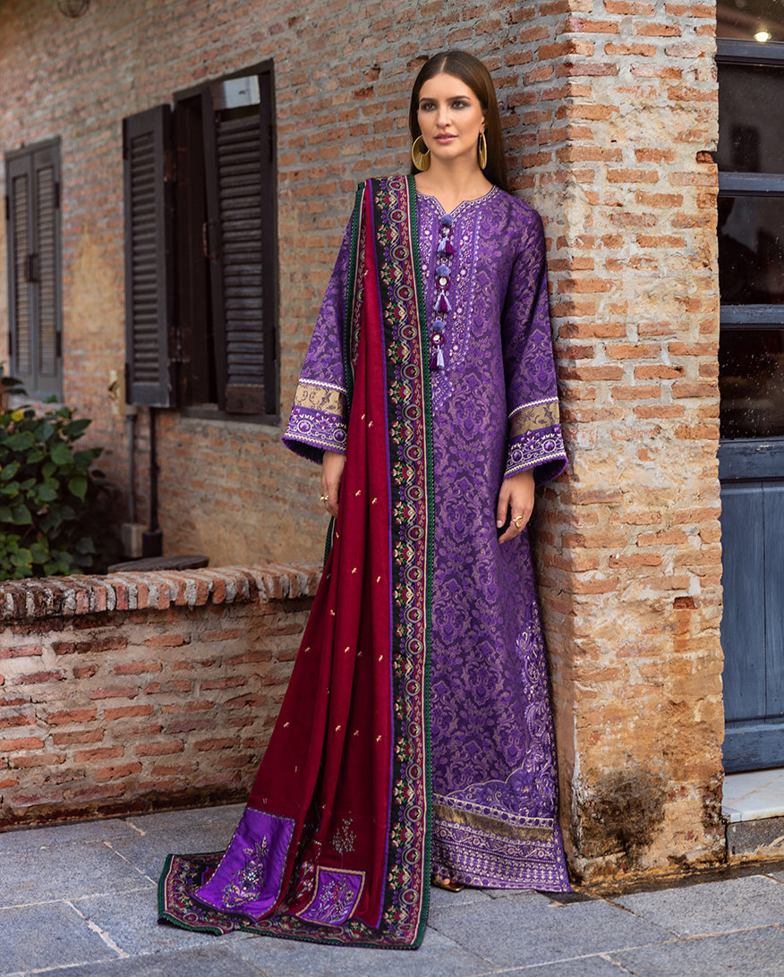 Woman wearing a deep purple jacquard full-length winter shirt with intricate gold-floral patterns and an embroidered contrasting crimson shawl standing outdoors near a brick wall.