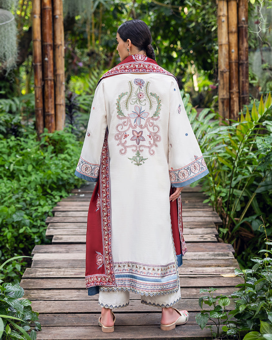 Rear view of woman in white embroidered Karandi suit with floral border designs and red shawl draped over shoulders, standing in natural garden setting