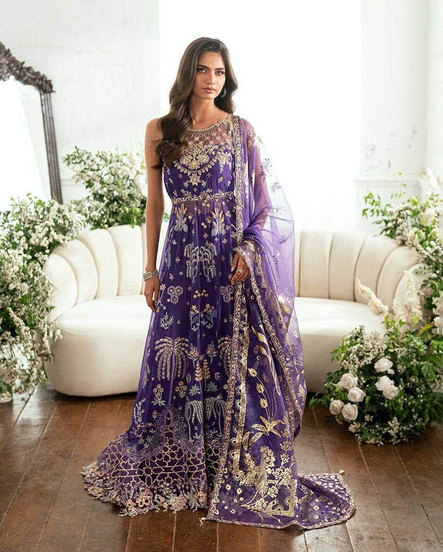 Front view of woman wearing a sleeveless purple chiffon gown with gold and pastel embroidery, holding a matching dupatta, standing in an elegant room with white floral decor