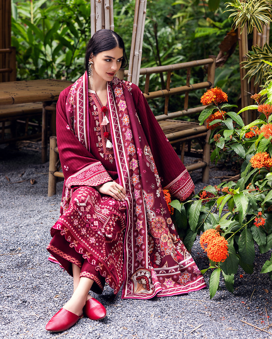 Seated woman wearing maroon embroidered karandi suit and matching shawl in outdoor garden setting