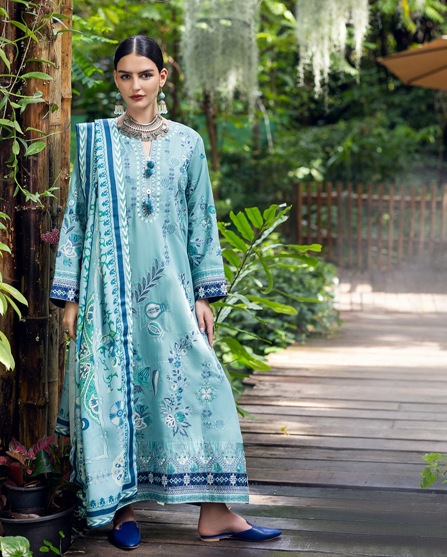 Full front view of the woman standing in a garden wearing the embroidered aqua blue karandi suit paired with a printed dupatta and blue slip-on shoes.