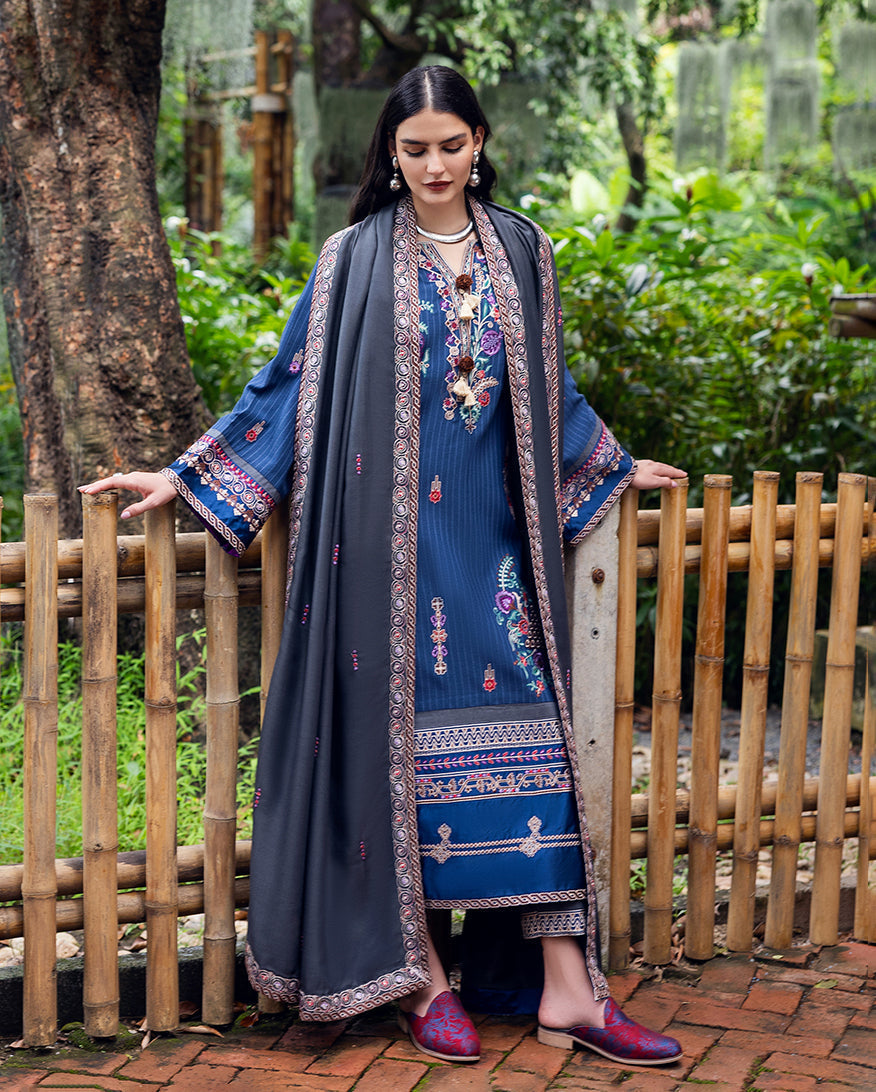 Front view of woman in full blue karandi suit with grey embroidered shawl holding bamboo fence in natural outdoor setting