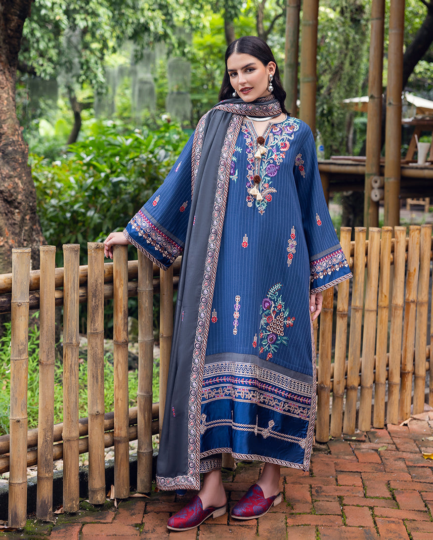 Woman wearing blue embroidered karandi suit with floral patterns and grey shawl standing by bamboo fence outdoors