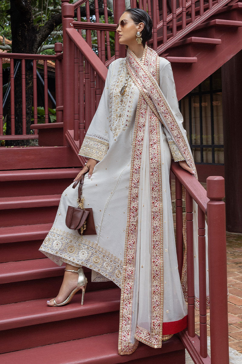Woman seated on red wooden stairs holding brown handbag, wearing embroidered ivory raw silk suit with gold high heels and embellished dupatta.