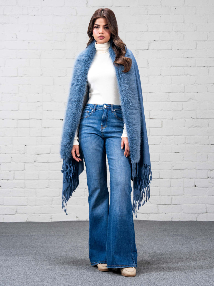 Woman wearing sky blue wool cape with faux fur trim and fringe over white turtleneck and blue flared jeans, standing against white brick wall, front view