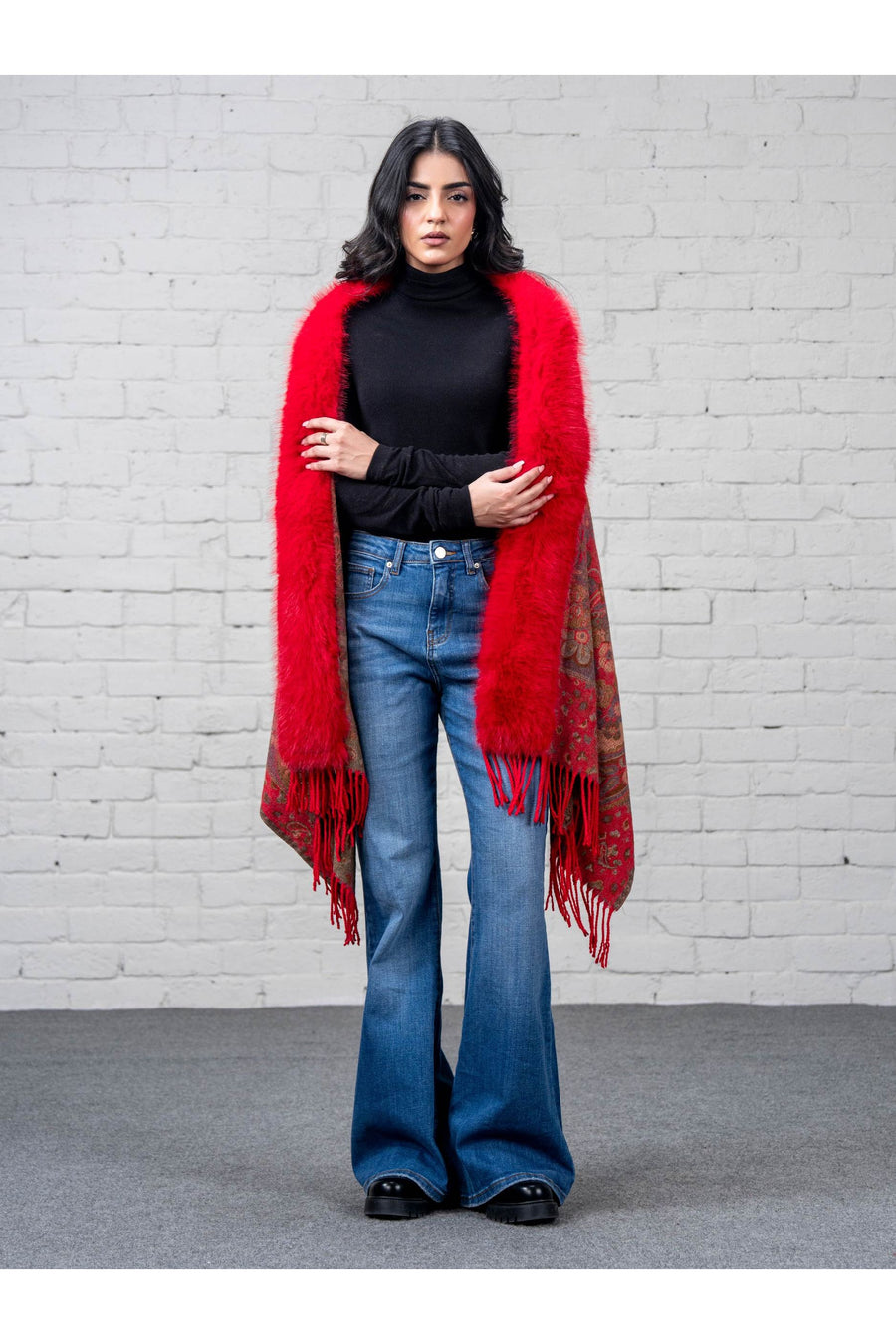 Front view of a woman wrapped in a detailed patterned merino wool cape with vivid red faux fur trim, styled with blue flared jeans and black turtleneck.
