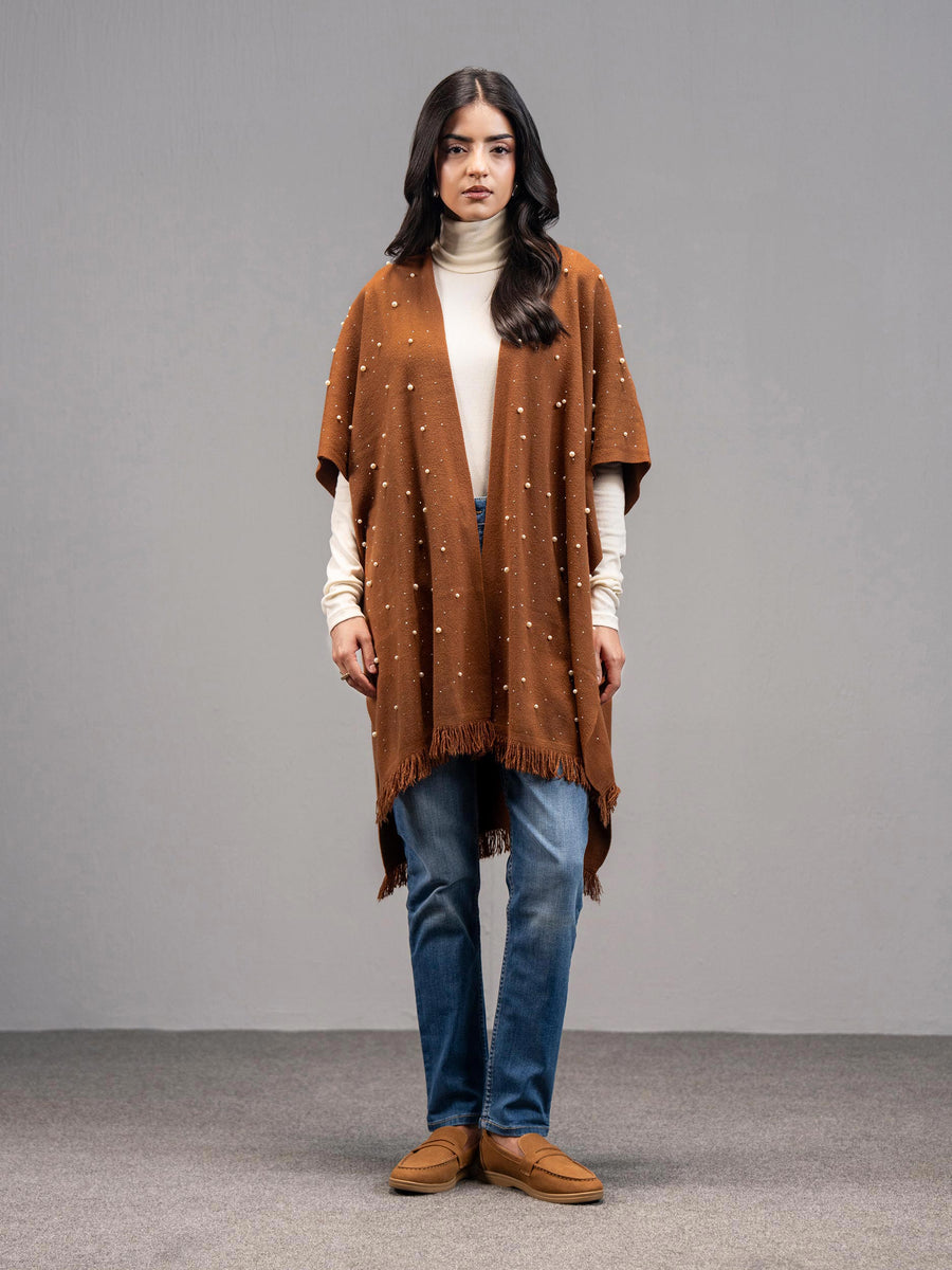 Front view of a woman wearing a rust-colored wool blend cape with pearl embellishments, paired with a white turtleneck and blue jeans.
