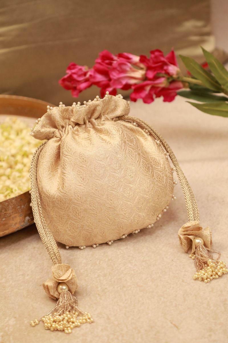 Ivory embroidered brocade potli bag with pearl bead tassels and drawstring closure on beige surface near pink flowers.