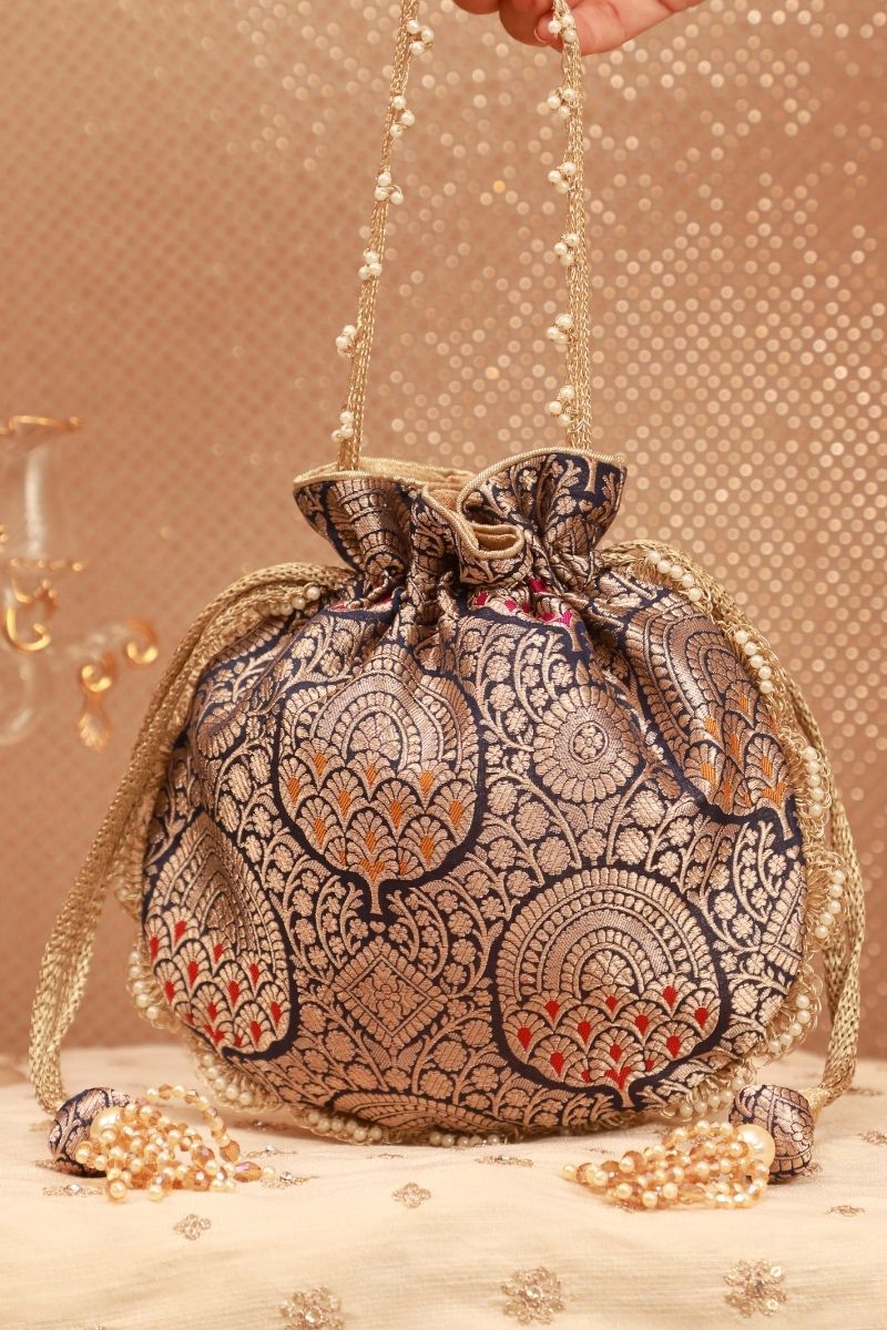 Navy blue Banarasi silk drawstring potli bag with intricate gold floral zari patterns, decorative gold trim, and beaded tassel accents held by hand.