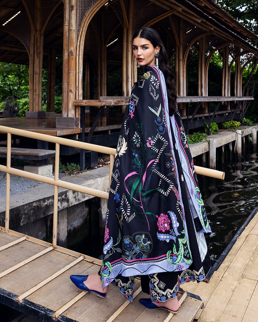 Back view of woman in black embroidered Karandi suit with printed shawl draped, standing outside near bamboo pillars and water