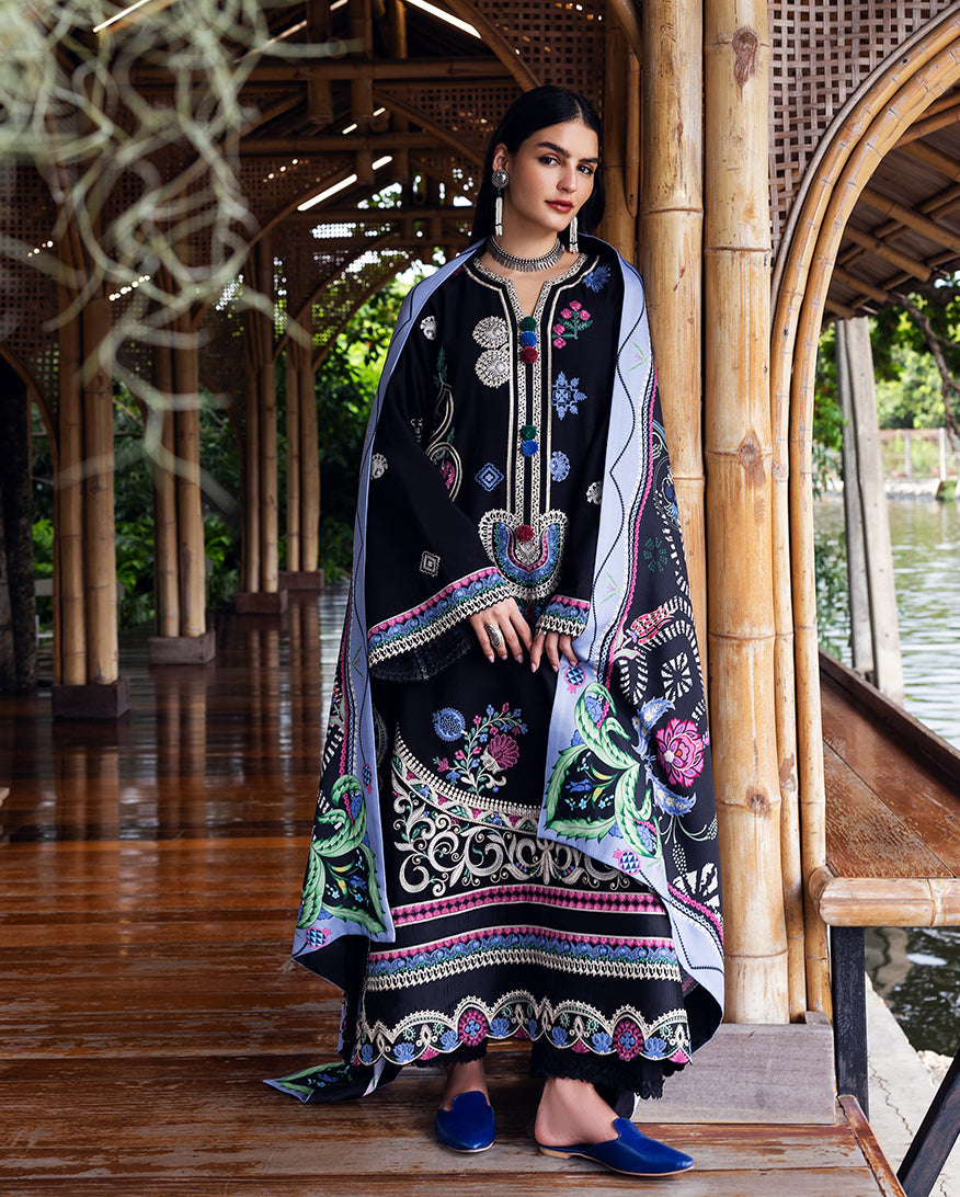 Woman seated wearing the three-piece Karandi suit including printed shawl and embroidered shirt, positioned next to bamboo pillars