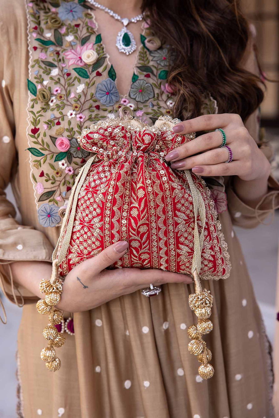 Close-up image of a woman holding a red embroidered potli bag with gold floral designs and gold tassels, wearing a beige dress with floral embroidery