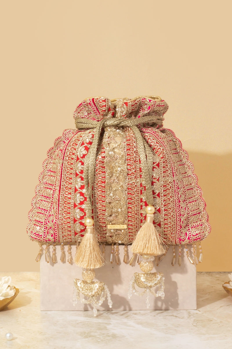 Red potli bag with detailed gold embroidery, tassels with bead and pearl accents, displayed on marble surface against a neutral background.