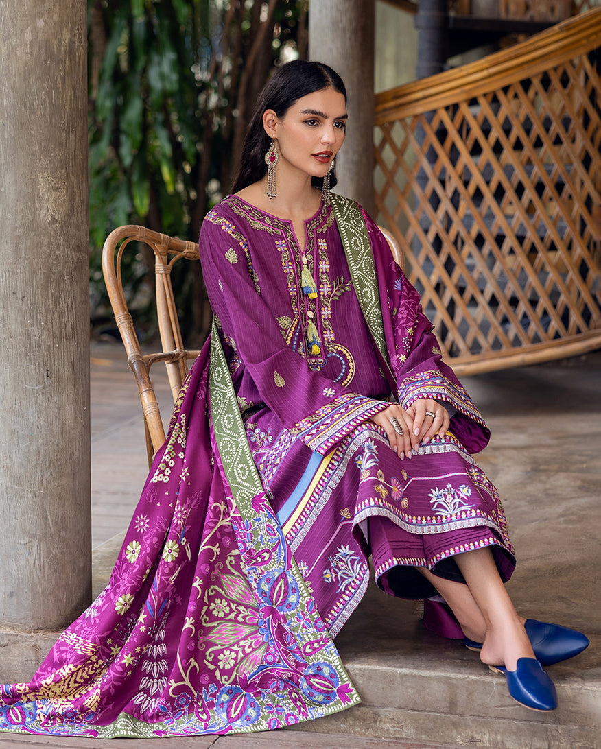 Seated model wearing rich plum purple embroidered Karandi suit paired with vibrant printed shawl and blue slip-on shoes.